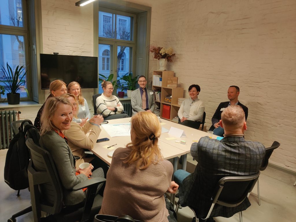 Smaidīga un pozitīva atmosfēra citā grupas diskusijā. Dalībnieki draudzīgi iesaistās sarunās un ideju apmaiņā pie galda. Telpas mājīgā vide un neformālais noskaņojums veicina atvērtu dialogu.