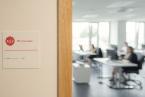 Pie durvīm redzama zīme ar numuru “422” un uzrakstu “Mācību klase”, kā arī teksts Braila rakstā. Fonā izplūdusi gaiša mācību telpa ar galdiem un cilvēkiem, kas strādā pie datoriem.