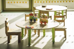 Gaiša pirmsskolas telpa ar nelieliem apaļiem galdiem un koka krēsliņiem. Uz galda novietotas koka rotaļlietas un krāsains pop-it, fonā redzami logi ar dabisku apgaismojumu un vēl viens bērnu galds.