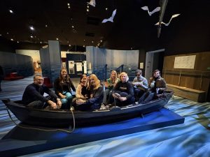 Projekta “Access Routes” partneri muzeja ekspozīcijā sēž vēsturiskā koka laivā, iepazīstot piekļūstamības un interpretācijas risinājumus. Telpā redzama jūras tematikas ekspozīcija ar instalācijām un informācijas stendiem.