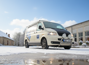 Balts mikroautobuss ar uzņēmuma logotipu stāv uz apsniguša ceļa ziemas dienā. Priekšējie lukturi ir ieslēgti, zem auto redzams atspīdums ledainā peļķē. Fonā – lauku ēkas un kaili koki zem zilām debesīm ar mākoņiem.