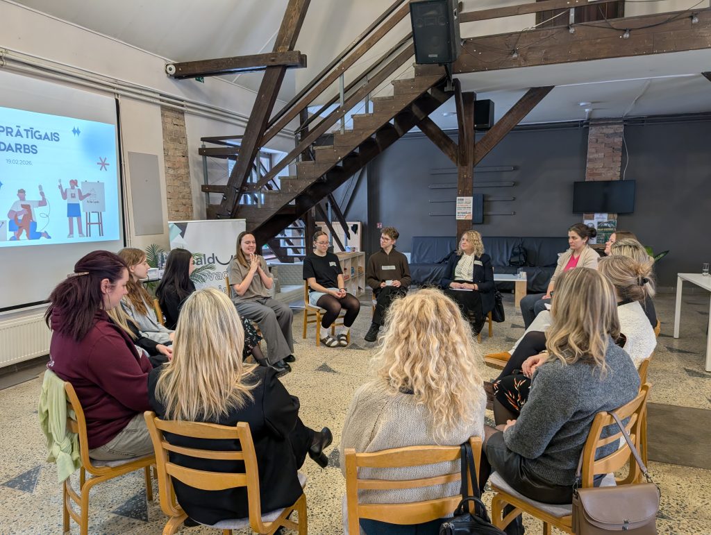 Plašā telpā ar koka sijām un kāpnēm dalībnieki sēž aplī uz krēsliem un piedalās diskusijā. Viena sieviete runā, bet pārējie klausās. Fonā redzams projekcijas ekrāns ar prezentāciju, dīvāns un atpūtas zona, radot semināra vai darbnīcas atmosfēru.