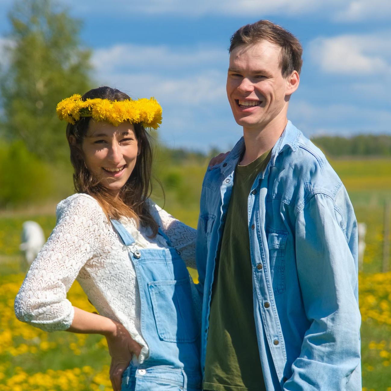 Jauns pāris stāv saulainā pļavā, kas pilna ar dzelteniem ziediem. Sievietei galvā ir pieneņu vainags, viņa smaida un tur roku uz gurna. Vīrietis stāv blakus un priecīgi smaida. Fonā redzama zaļa ainava un zilas debesis ar mākoņiem.