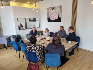 Foto redzama darba tikšanās nelielā konferenču telpā – pie galda sēž seši dalībnieki ar klēpjdatoriem un dokumentiem, notiek diskusija. Uz galda kafijas krūzes un pieraksti, fonā gleznas un moderns interjers.