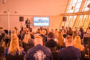 Konferences dalībnieki klausās prezentāciju Liepājas koncertzālē “Lielais dzintars”. Priekšā uz skatuves runātājs pie ekrāna, kur redzams prezentācijas slaids, bet zālē sēž dažādu nozaru tūrisma profesionāļi.