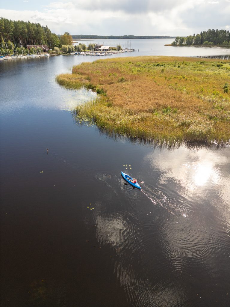 Skats no augšas uz mierīgu ezeru, kurā pa tumšo ūdeni airē viens cilvēks zilā kajakā, atstājot vieglas ripples. Krastā redzama niedru josla un neliela jahtu piestātne ar laivām, bet ūdens virsmā atspoguļojas mākoņi.