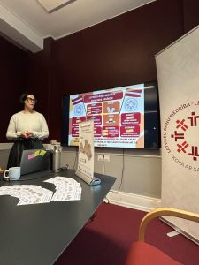 Sieviete prezentē informāciju par latviešu diasporas aktivitātēm Dānijā, stāvot pie ekrāna ar pasākumu kalendāru. Uz galda izvietoti informatīvie materiāli par remigrācijas iespējām Latvijā, veicinot sadarbību ar tautiešiem ārvalstīs.