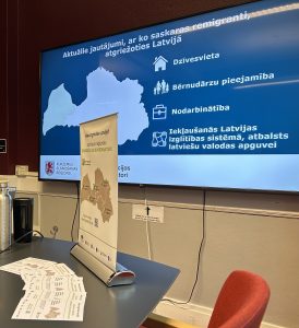 Prezentācijas slaids par aktuālajiem jautājumiem, ar kuriem saskaras remigranti, atgriežoties Latvijā. Ekrānā izcelti būtiskākie aspekti – dzīvesvieta, bērnudārzu pieejamība, nodarbinātība un iekļaušanās izglītības sistēmā. Uz galda redzami informatīvie materiāli par remigrācijas iespējām.