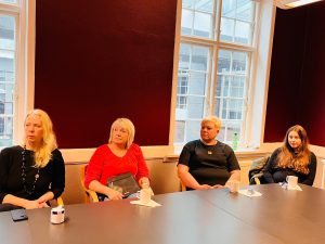 Četras sievietes sēž pie galda tikšanās laikā, klausoties prezentāciju par remigrācijas iespējām. Uz galda redzamas kafijas krūzes un piezīmes, telpā valda mierīga, koncentrēta atmosfēra, veicinot diskusiju ar diasporas pārstāvjiem.