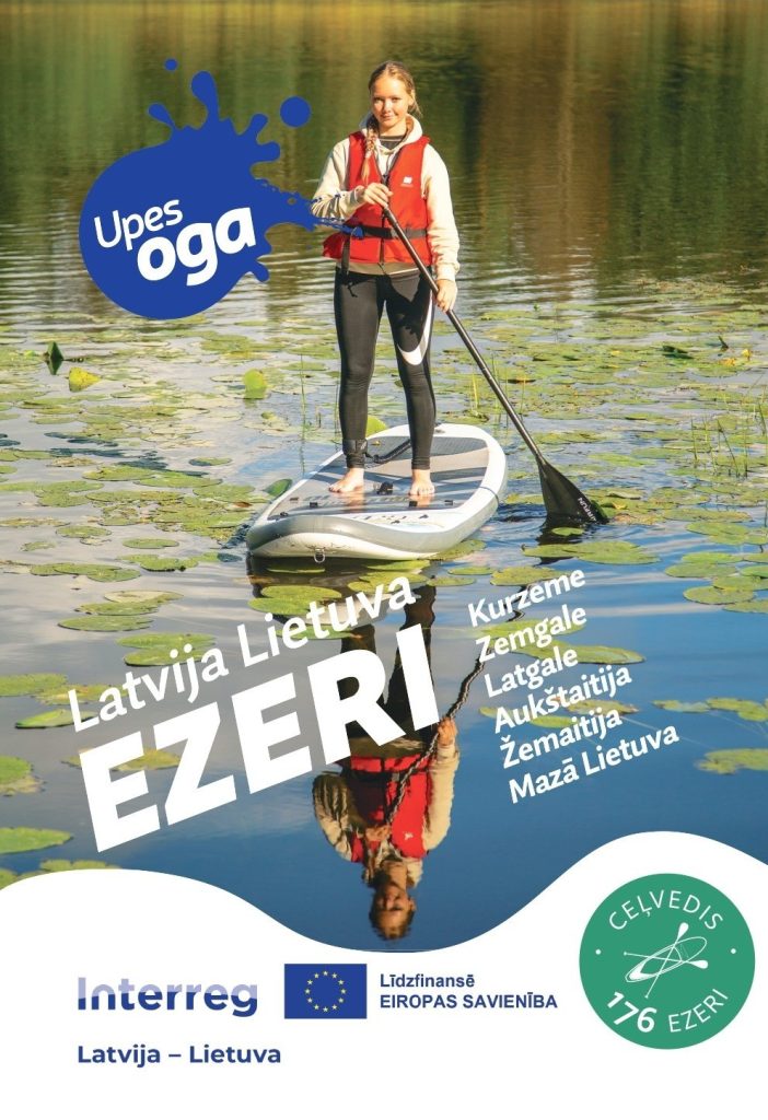 Meitene ar glābšanas vesti stāv uz SUP dēļa ezera ūdenī, apkārt peld ūdensrozes. Uz attēla redzams “Upesoga” logotips un uzraksts “EZERI Latvija Lietuva”, kā arī norādes uz reģioniem un Interreg atbalstu.