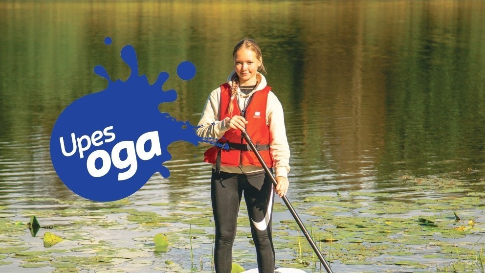 Meitene ar glābšanas vesti stāv uz SUP dēļa mierīgā ezerā, ko klāj ūdensrozes. Attēlā redzams “Upesoga” logotips, bet fonā – saulaina dabas ainava ar mierīgu ūdens virsmu un krasta atspulgu.