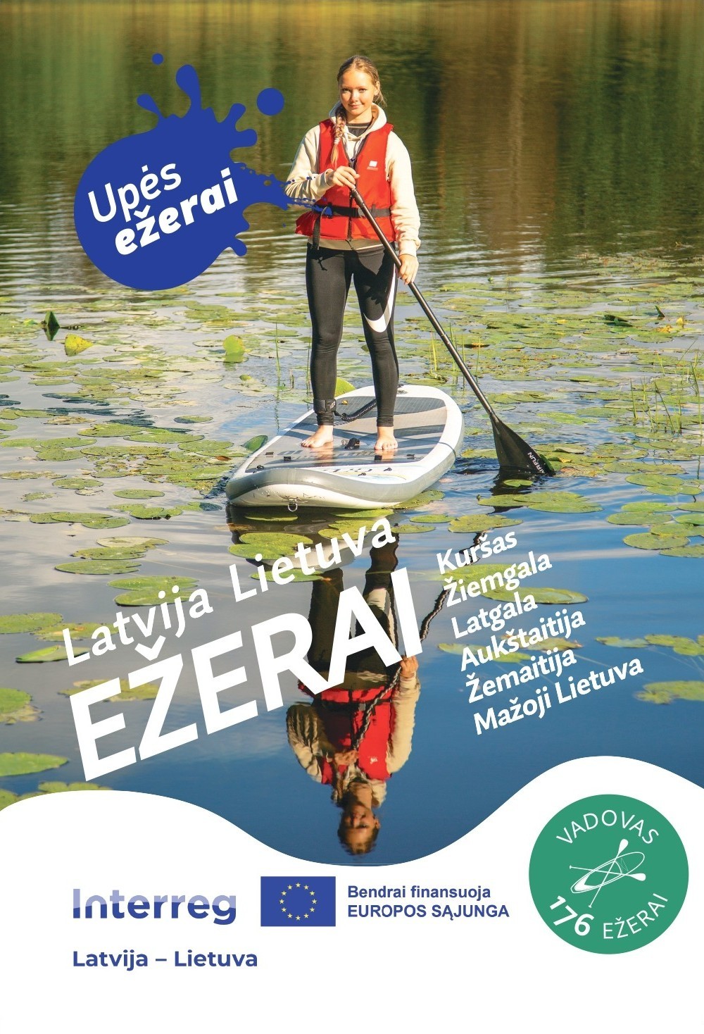 Meitene ar glābšanas vesti stāv uz SUP dēļa ezera ūdenī, apkārt ūdensrozes. Attēlā redzams uzraksts “EZERAI Latvija Lietuva” lietuviešu valodā, reģionu nosaukumi un Interreg programmas līdzfinansējuma norāde.