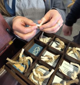 Cilvēks rokās tur virvju mezglu un pēta to no ceļojošā muzeja lādes nodalījuma. Koka kastē redzami dažādi mezgli ar aprakstiem un vizuāliem attēliem, kas ļauj apmeklētājiem izzināt jūrniecības tematu caur tausti un praktisku pieredzi.