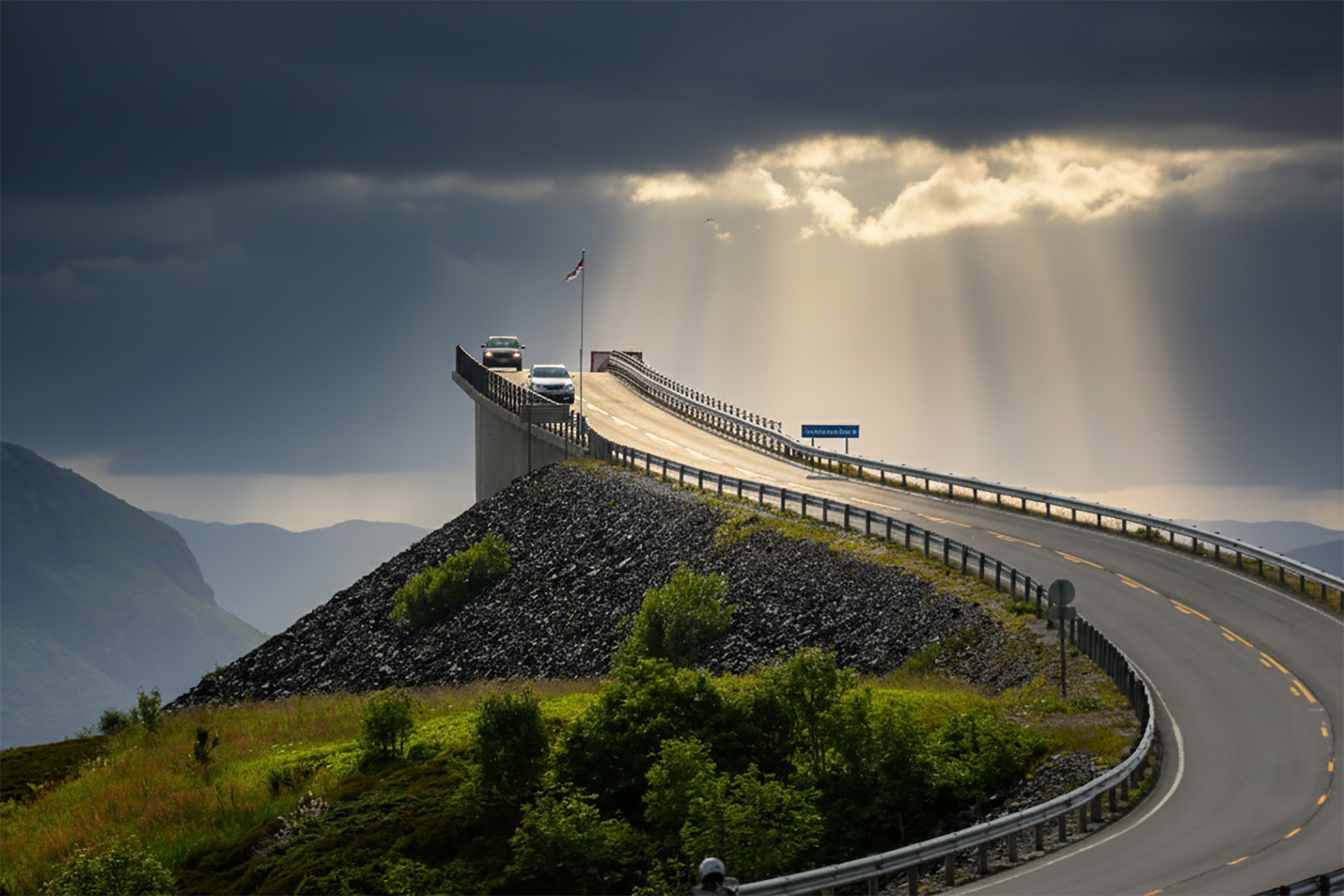 Kalnu ceļš ar asu līkumu ved pāri nelielam uzbērumam, pa kuru brauc automašīnas. Virs horizonta starp tumšiem mākoņiem laužas saules stari, izgaismojot ceļa posmu un apkārtējo ainavu ar zaļiem krūmiem un kalnu reljefu.