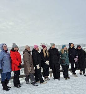 Projekta partneri ziemas pārgājiena laikā stāv uz skatu platformas sniegotā mežā Somijā. Dalībnieki siltā ziemas apģērbā vēro apkārtējo ainavu ar sniegu klātiem kokiem un paugurainu mežu fonā.