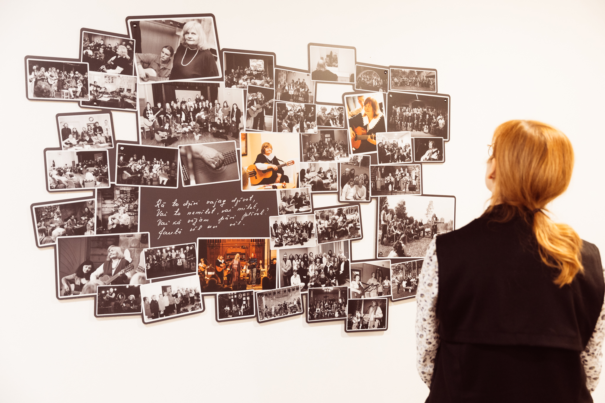 Apmeklētāja aplūko sienas instalāciju, kurā izvietotas dažādas melnbaltas un krāsainas fotogrāfijas no muzikālām un kultūras norisēm, veidojot vizuālu stāstu par cilvēkiem, koncertiem un radošo darbību.