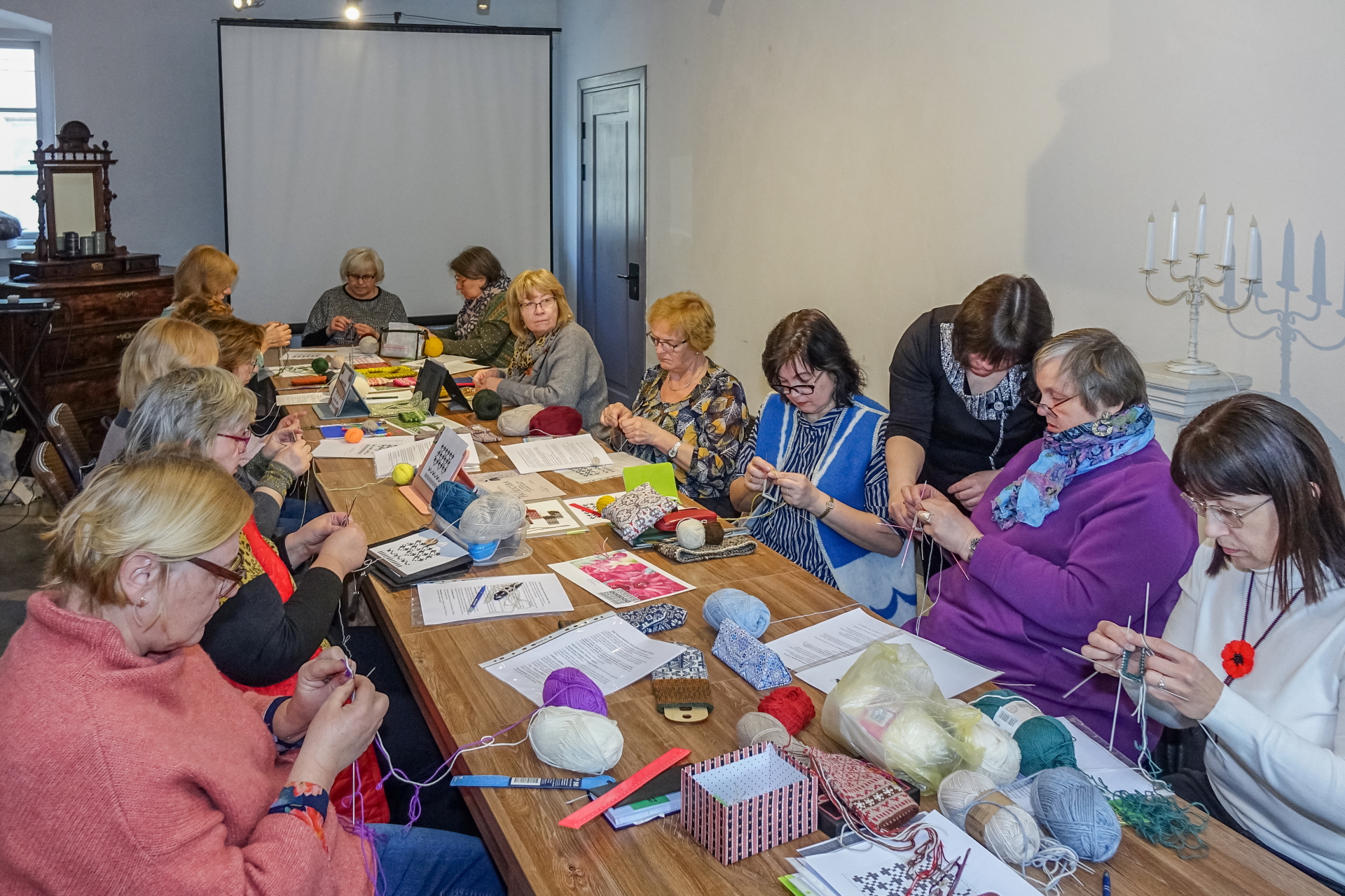 Amatu mājā pie gara koka galda pulcējušies dažāda vecuma dalībnieki, kas kopīgi ada ar krāsainām dzijām. Uz galda redzami materiāli, adāmadatas un instrukcijas, valda mierīga, radoša darbnīcas atmosfēra.