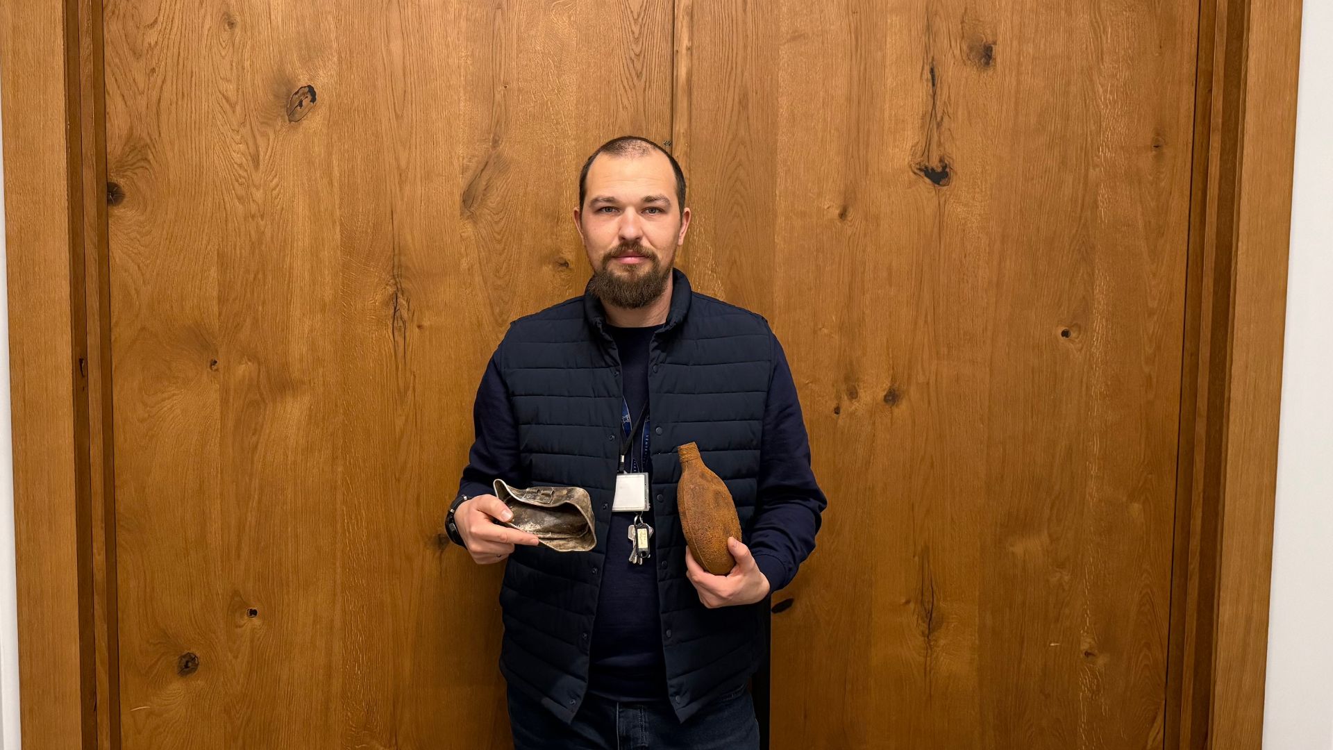 Vīrietis stāv pie koka sienas un rokās tur divus arheoloģiskus priekšmetus – metāla trauka fragmentu un senas pudeles formu. Attēls uzņemts iekštelpās, dokumentējot vēstures vai muzeja aktivitāti.