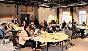 Plašā, modernā sanāksmju telpā pie apaļiem galdiem sēž dalībnieki ar datoriem un dokumentiem, klausoties prezentāciju. Telpā redzamas ķieģeļu sienas, projektors un tāfele, radot profesionālu darba semināra atmosfēru.