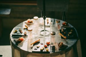 Apaļš galds ar baltu galdautu, uz kura izkārtotas nelielas degustācijas uzkodas uz tumšām un gaišām šķīvju plāksnēm. Apkārt novietotas glāzes un mazas pudelītes ar dzērieniem, galdu apspīd silta saules gaisma, radot mierīgu un izsmalcinātu maltītes atmosfēru.