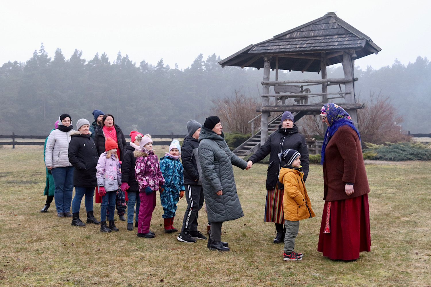 Bērni un pieaugušie rindā pļavā Jūrkalnē satiekas ar sievieti tautastērpā, simboliski sveicinot pavasari pavasara saulgriežu tradīcijās. Fonā redzams koka skatu tornis un miglains piejūras ainavas fons.
