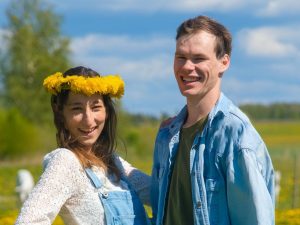 Jauns pāris stāv saulainā pļavā, kas pilna ar dzelteniem ziediem. Sievietei galvā ir pieneņu vainags, viņa smaida un tur roku uz gurna. Vīrietis stāv blakus un priecīgi smaida. Fonā redzama zaļa ainava un zilas debesis ar mākoņiem.