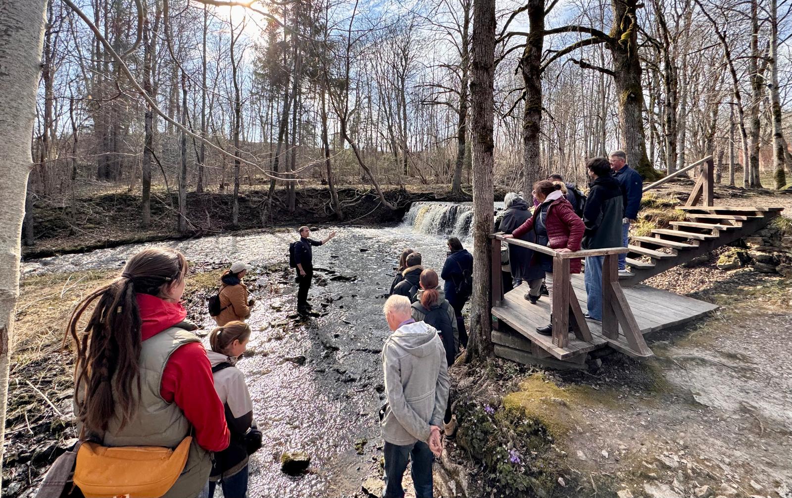 Apmācību dalībnieki pulcējušies pie Abavas ūdenskrituma, kur pasniedzējs skaidro ūdens erozijas procesus un reljefa veidošanos, vērojot dabas norises klātienē.