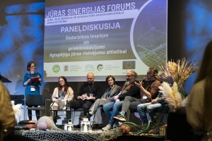 Paneļdiskusijā eksperti diskutē par sadarbības iespējām un priekšnosacījumiem ilgtspējīgu jūras risinājumu attīstībai. Uz skatuves redzami dažādu nozaru pārstāvji, auditorija seko līdzi diskusijai.