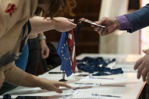 Foruma reģistrācijas laikā dalībniekam tiek izsniegta identifikācijas karte un materiāli. Uz galda redzamas Eiropas Savienības un Latvijas karodziņi, kas simbolizē starptautisko sadarbību un pasākuma kontekstu.