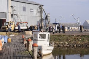 Foruma dalībnieki pulcējušies Liepājas ostā pie industriālās teritorijas, iepazīstot piekrastes zvejniecības vidi un infrastruktūru. Priekšplānā redzama pietauvota laiva, bet fonā – ostas celtņi, kas raksturo zilās ekonomikas un jūras nozaru attīstību.
