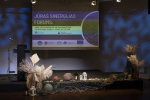 Uz skatuves redzams Jūras sinerģijas foruma prezentācijas ekrāns ar pasākuma nosaukumu un tematiku. Priekšplānā veidota jūras tematikai atbilstoša scenogrāfija ar zvejas tīkliem, bojām un dabas elementiem.