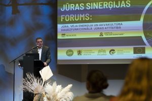 Foruma atklāšanas laikā uz skatuves uzstājas runātājs, prezentējot Jūras sinerģijas foruma tematiku. Fonā redzams pasākuma vizuālais noformējums un partneru logotipi, auditorijā – klausītāji.