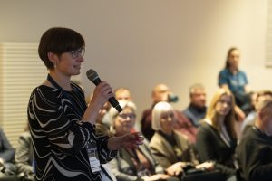 Diskusiju laikā foruma dalībniece uzdod jautājumu, izmantojot mikrofonu, kamēr pārējie klausītāji uzmanīgi seko līdzi. Attēls atspoguļo aktīvu iesaisti un viedokļu apmaiņu zilās ekonomikas jautājumos.