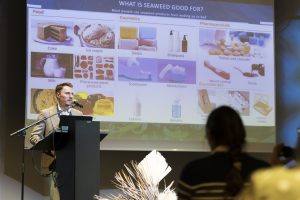 Prezentācijas laikā runātājs skaidro jūras resursu, īpaši aļģu, izmantošanas iespējas pārtikā, kosmētikā un farmācijā. Fonā redzams informatīvs slaids, auditorijā – klausītāji.