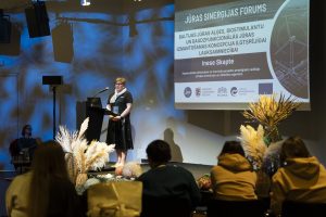 Prezentācijas laikā runātāja iepazīstina ar Baltijas jūras aļģu un biostimulantu izmantošanu ilgtspējīgā lauksaimniecībā. Fonā redzams foruma vizuālais noformējums, auditorijā – klausītāji.