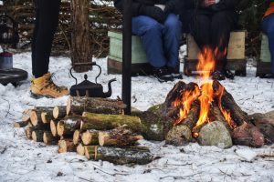 Cilvēki sēž ap ugunskuru sniegotā mežā, sildoties pie liesmām. Blakus sakrauta malka un novietota metāla tējkanna, radot mierīgu, dabisku un kopības pilnu atmosfēru ārpus telpām.