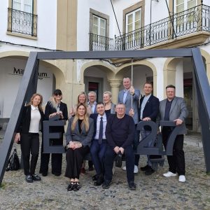 Grupas foto ar semināra dalībniekiem pie viesnīcas Pullman Rīgā. Dalībnieki pozē pie instalācijas ar uzrakstu “ELA 27”, fonā redzama ēkas fasāde ar arkām un balkoniem. Attēlā redzami dažādu institūciju pārstāvji.