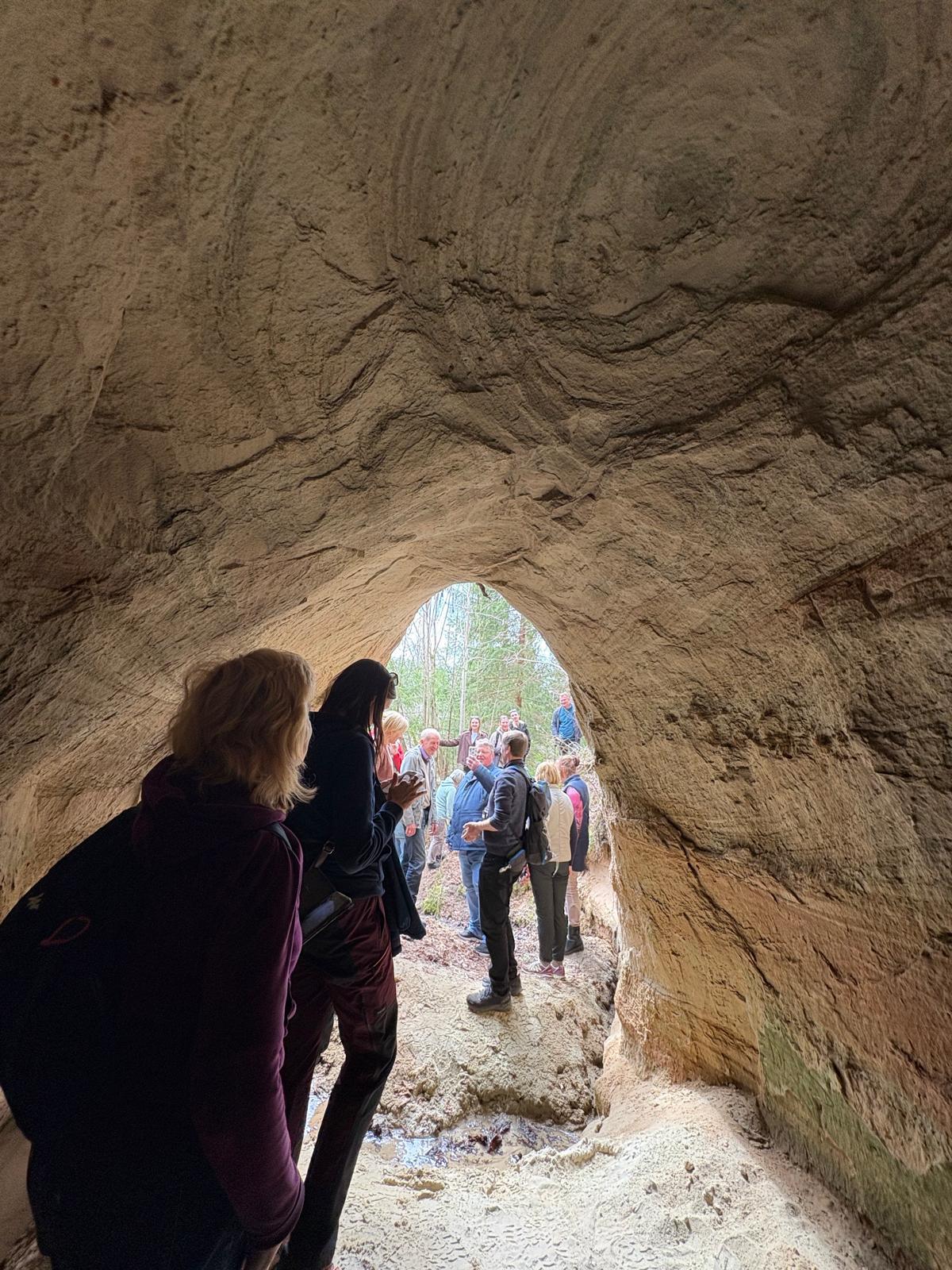 Apmācību dalībnieki atrodas smilšakmens alā Abavas ielejā, klausoties gida stāstījumā par ģeoloģiskajiem procesiem. Caur alas atveri redzama daba un pārējā grupa ārpusē.