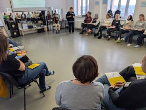 Plašā telpā cilvēki sēž aplī un piedalās diskusijā vai apmācībās. Vadītāja stāv vidū un uzrunā dalībniekus, kuri klausās un veic piezīmes. Redzami prezentācijas materiāli un darba burtnīcas.