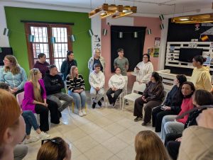 Telpā cilvēki sēž aplī un piedalās diskusijā vai grupu darbā. Viens dalībnieks runā, kamēr pārējie klausās, veidojot iesaistošu un sadarbībā balstītu mācību vidi.