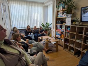 Mājīgā telpā dalībnieki sēž uz dīvāna un krēsliem, skatās prezentāciju ekrānā un klausās. Redzama neformāla mācību vide ar kafiju, uzkodām un plauktiem ar spēlēm un grāmatām.