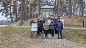 Apmeklētāja skenē QR kodu uz soliņa dabas teritorijā, izmantojot viedtālruni, kamēr fonā citi dalībnieki pārvietojas pa mežainu apkārtni pie zaļas koka ēkas. Attēls ilustrē digitālos risinājumus un pieejamības uzlabojumus pārgājienu maršrutos.