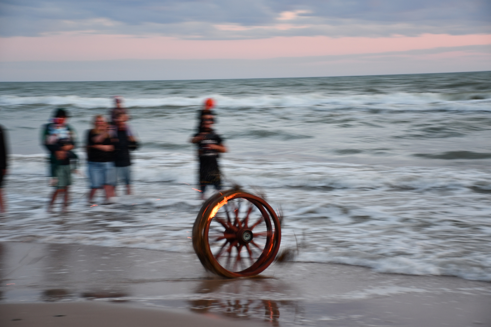 Jūras krastā notiek uguns rituāls – deg ritenis tiek ripināts viļņos, fonā redzami cilvēki, kas vēro notiekošo. Attēlā iemūžināta dinamiska un simboliska Saulgriežu tradīcija pie jūras.