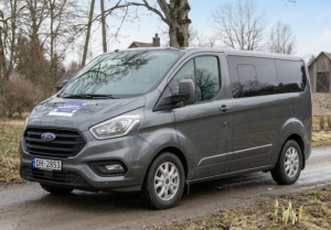 Pelēks mikroautobuss no Vānes transporta pēc pieprasījuma pilotteritorijas brauc pa lauku ceļu Kurzemē. Transportlīdzeklis nodrošina iedzīvotāju mobilitāti, savienojot apdzīvotas vietas ar tuvākajiem pakalpojumu centriem.