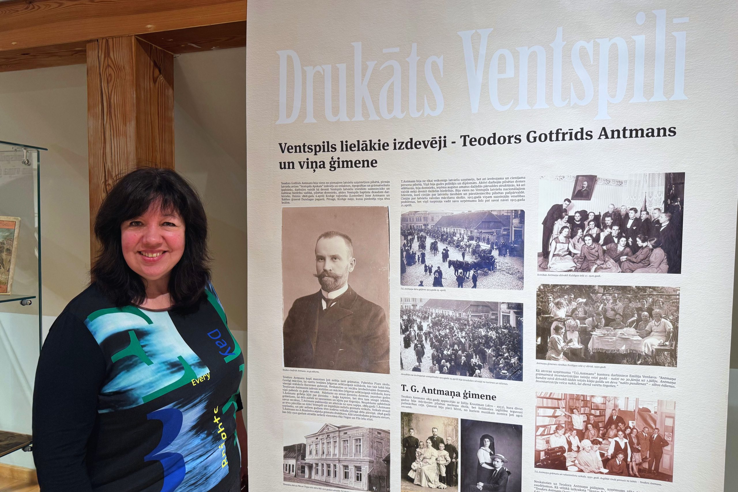 Ventspils muzeja vadošā vēsturniece Māra Dāvida stāv pie ekspozīcijas stenda “Drukāts Ventspilī”, kas veltīts izdevējam Teodoram Gotfrīdam Antmanam un viņa ģimenei. Redzamas vēsturiskas fotogrāfijas un apraksti.