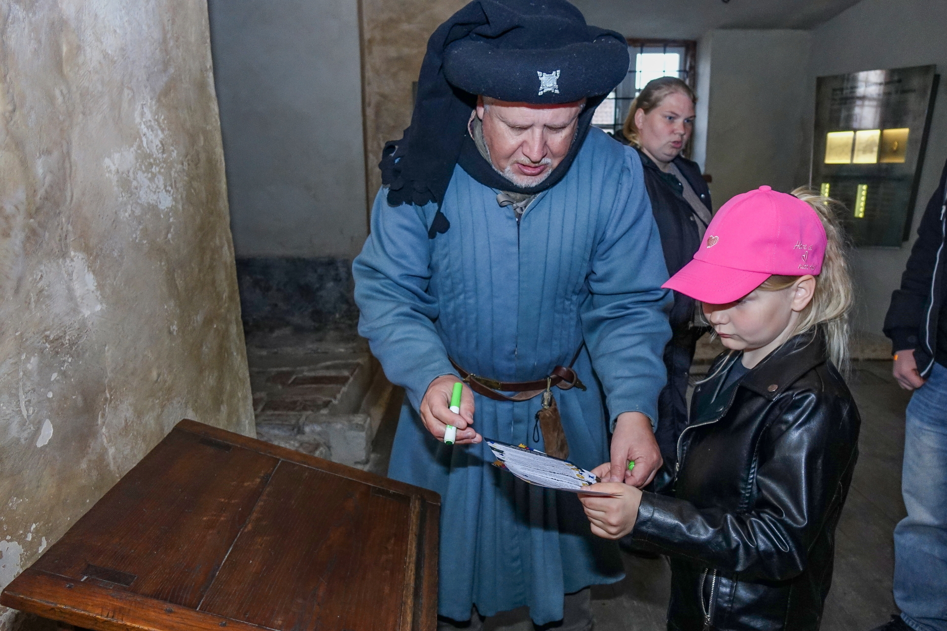 Muzeju nakts pasākumā Ventspils muzejā vēstures tēlā ģērbts gids palīdz bērnam aizpildīt uzdevumu lapu. Attēlā redzama izzinoša aktivitāte muzeja telpās, kur apmeklētāji iesaistās vēstures izzināšanā.