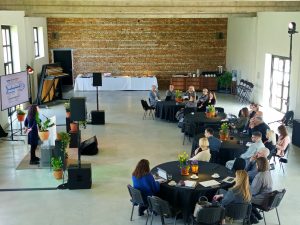 Seminārs koncertzālē “DUNA” Kuldīgā – dalībnieki pie apaļajiem galdiem klausās prezentāciju par energopārvaldību. Plašajā telpā redzama skatuve, ekrāns ar vizuālajiem materiāliem un pavasarīgi dekorēti galdi.