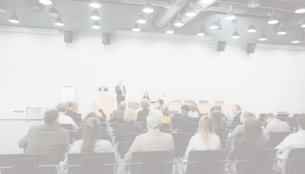 Semināra telpa ar auditoriju – cilvēki sēž rindās un klausās prezentāciju. Priekšā runātājs uzstājas pie pults, fonā ekrāns un paneļdiskusijas dalībnieki. Mūsdienīga konferenču vide ar apgaismojumu un tehnisko aprīkojumu.