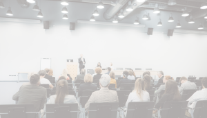 Semināra telpa ar auditoriju – cilvēki sēž rindās un klausās prezentāciju. Priekšā runātājs uzstājas pie pults, fonā ekrāns un paneļdiskusijas dalībnieki. Mūsdienīga konferenču vide ar apgaismojumu un tehnisko aprīkojumu.