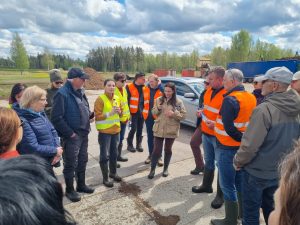 Cilvēku grupa pulcējas apļveida sarunu lokā; vairāki dalībnieki ģērbti oranžās un dzeltenzaļās atstarojošās vestēs. Fonā redzama celtniecības tehnika, zemes kaudzes un zils konteiners.